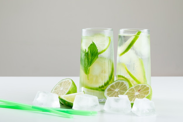 Free photo lemonade in glasses with lemons, straws, basil, ice cubes side view on white and grey