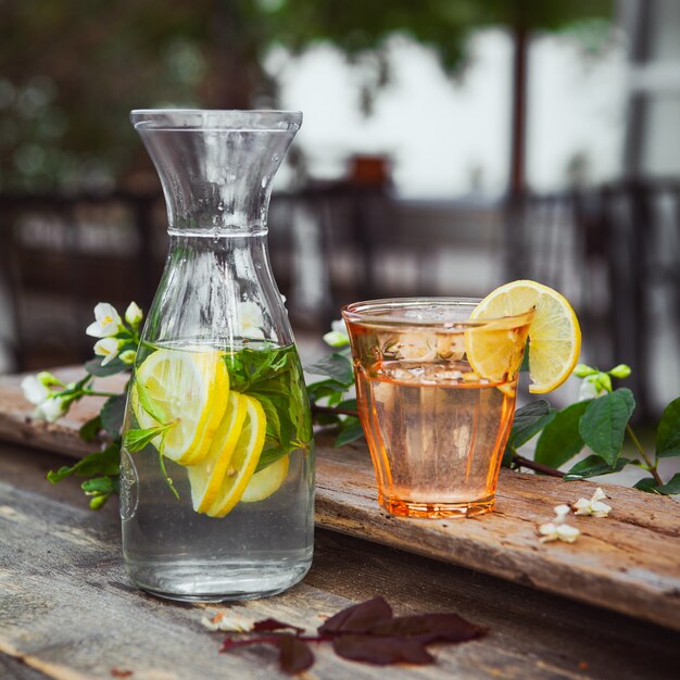 Lemonade with flowers on branch in glass and jug on wooden and yard table, side view.