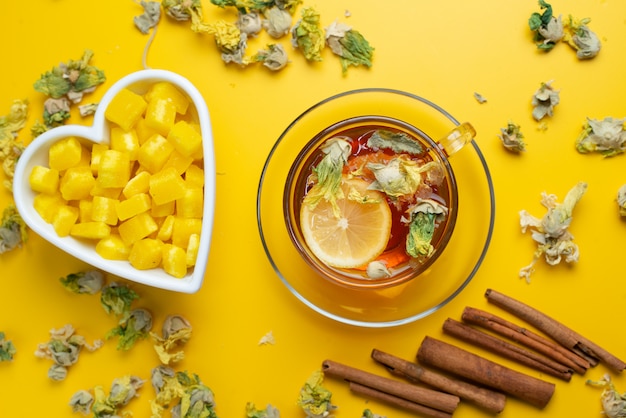 Lemony tea with dried herbs, sugar cubes, cinnamon sticks in a cup on yellow surface, flat lay.