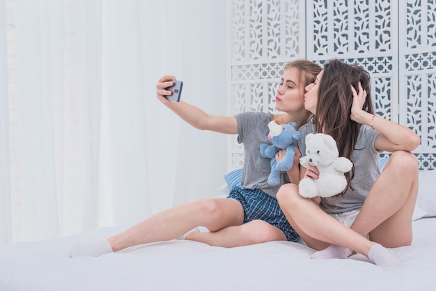 Lesbian couple sitting on bed holding soft toys taking selfie on mobile phone