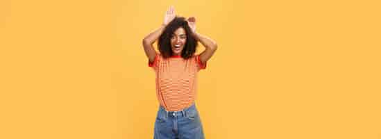 Free photo let me be your bunny portrait of charming enthusiastic and charismatic happy darkskinned female with afro hairstyle holding palms on head like animal ears making cute face over orange background