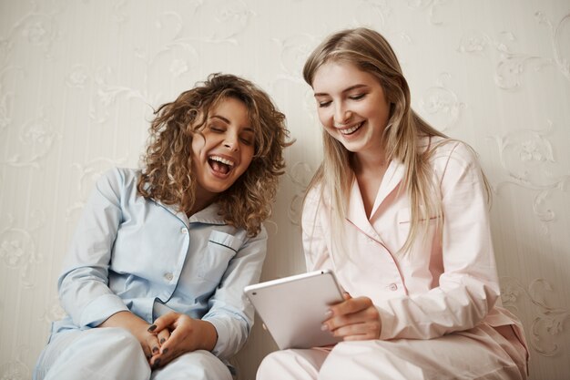 Let me show you funny video. Portrait of beautiful caucasian blond sister in nightwear spending leisure with friend, holding digital tablet while reading hilarious joke or article, having fun together