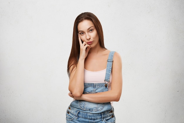 Let me think. Smart cunning quickwitted beautiful gir in jeans jumpsuit looking pensive while plotting or scheming something, holding hand on her cheek.