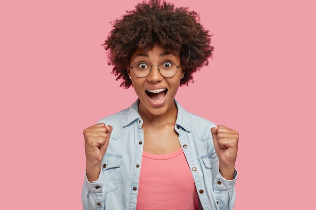 Lets celebrate my success! Attractive curly dark skinned female raises clenched fists, rejoices triumph or win, wears round spectacles and jean jacket, feels thrilled and amused, isolated on pink wall