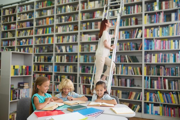 Library. Joyful primary school children reading at table and young long-haired woman on stairs near bookshelf in library