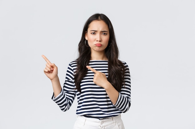 Lifestyle, beauty and fashion, people emotions concept. Disappointed sad asian girl complaining, feeling regret or jealousy as pouting uneasy and pointing fingers left at promo banner