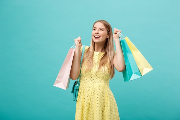 Lifestyle Concept: Portrait of shocked young attractive woman in yellow summer dressposing with shopping bags and looking at camera over blue background.