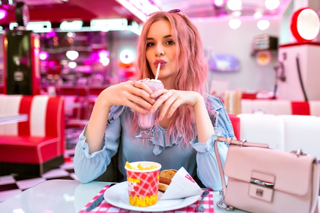 Lifestyle indoor image of stylish young pretty woman with wavy unusual pink hairs and natural make up, wearing cute blue dress and denim jacket, enjoy her tasty American dinner.