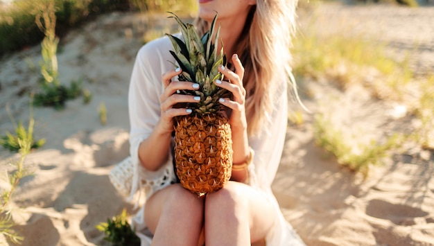 Lifestyle outdoor picture of laughing pretty woman with juicy pineapple relaxing on sunny beach . Trendy summer outfit