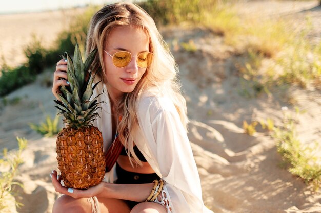 Lifestyle outdoor picture of laughing pretty woman with juicy pineapple relaxing on sunny beach . Trendy summer outfit