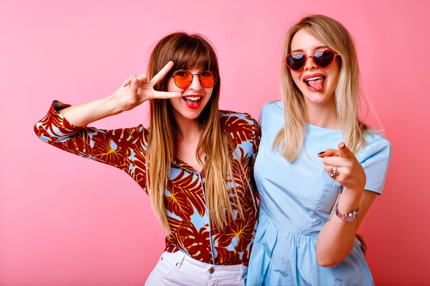 lifestyle portrait of happy pretty two best friends sister girls, posing and having fun together at pink wall,showing long tongue and v gesture, positive party mood.