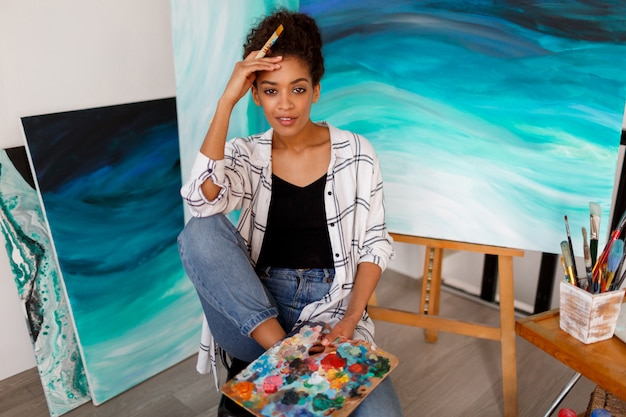 Free photo lifestyle portrait  of  young african student sitting with amazing abstract sea acrylic hand drawn artwork at the studio.