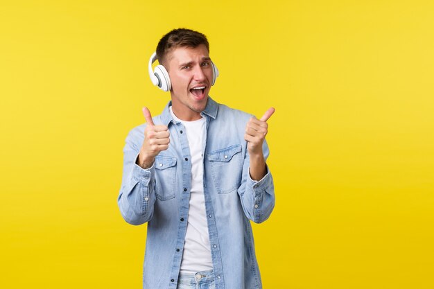 Lifestyle, summer holidays, technology concept. Excited handsome happy blond man vibing, chilling with awesome music, wearing headphones and showing thumbs-up in approval, yellow background.