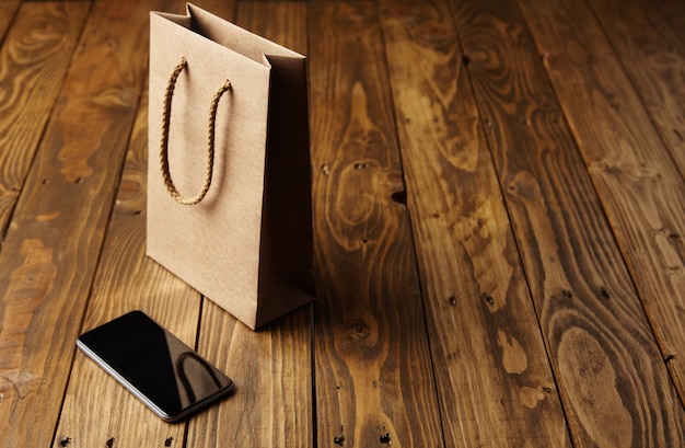 Free photo light brown craft paper bag reflecting in a spotless black smartphone lying next to it on a handcrafted wooden table