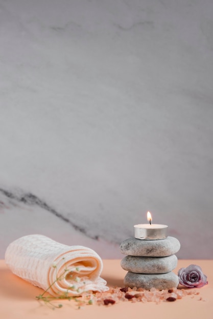 Lighted candle over the spa stones with napkin; rose and himalayan salts on peach colored backdrop against grey background