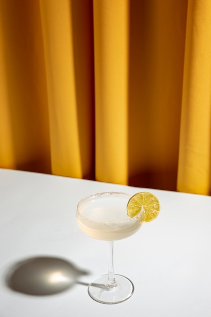 Lime cocktail in a champagne saucer on white desk against yellow curtain