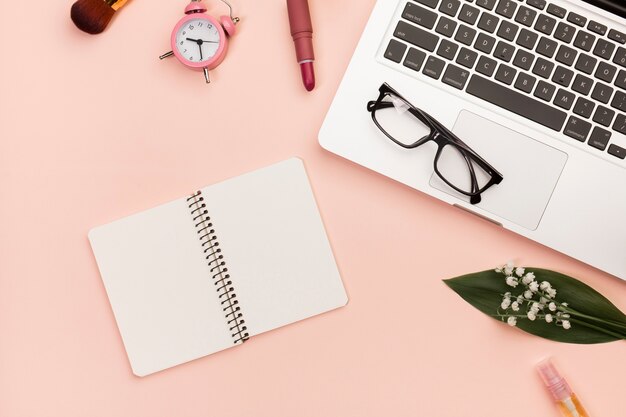 Lipstick,makeup brush,alarm clock,eyeglasses on laptop,spiral notepad on peach background