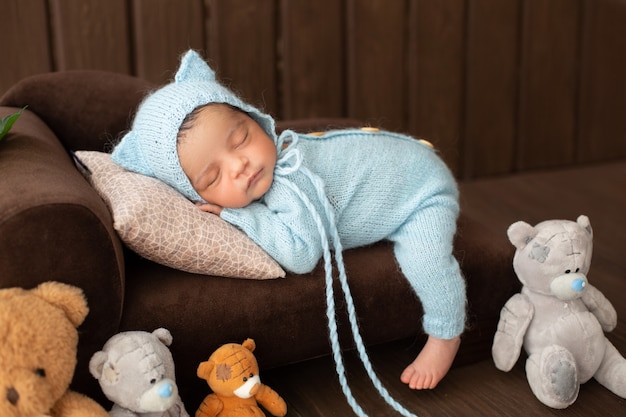 Little baby boy pretty newborn infant laying on brown sofa in blue crocheted pijama surrounded by toys