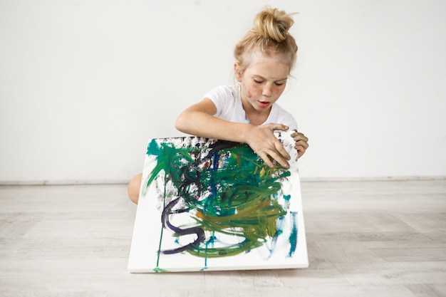 Free Photo little blonde female child with hairbn and freckles wearing white t-shirt occupied with her picture. cute, adorable girl sitting on a floor with colourful canvas on her knees.