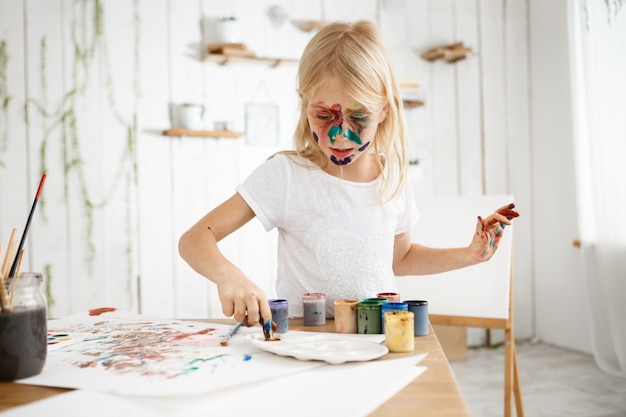 Free photo little blonde girl busy and concentrated on mixing paint on palette.