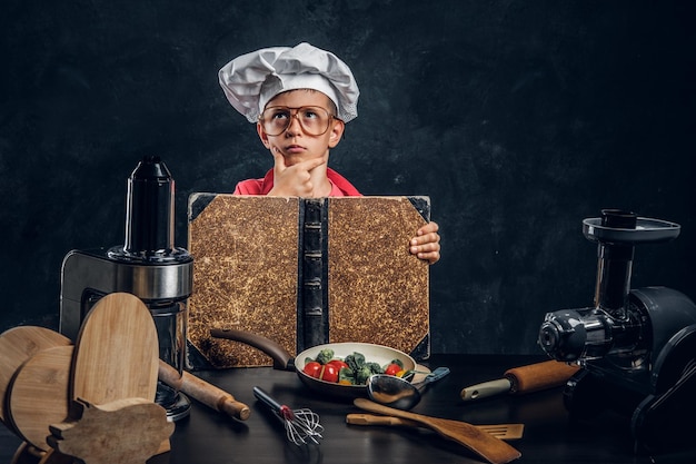 Little boy in glasses and chef's hat is reading recipe book and planing to cook.