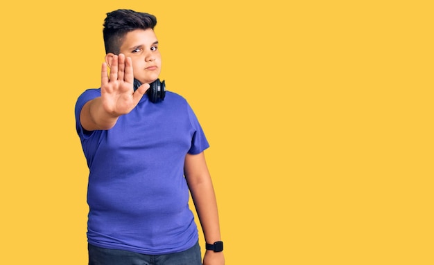 Little boy kid listening to music wearing headphones doing stop sing with palm of the hand. warning expression with negative and serious gesture on the face.