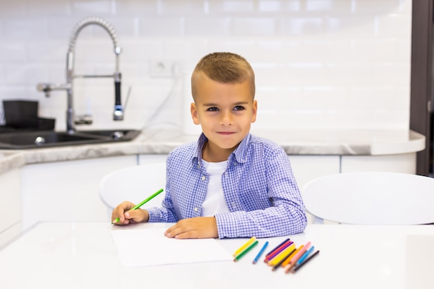 Little boy sits at a table in a bright kitchen and draws with pencils