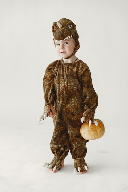 Free photo little boy toddler dressed in brown costume of a dinosaur holding a pumpkin. boy has a hood with dino's face