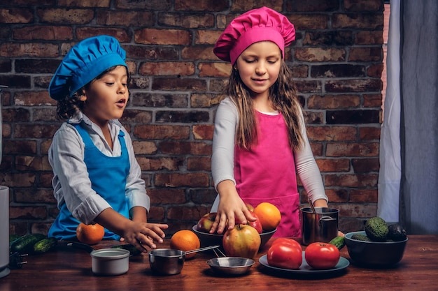 Free photo little boy with brown curly hair dressed in a blue cook uniform and a beautiful girl dressed in a pink cook uniform cooking together in a kitchen against a brick wall. cute little cook couple.