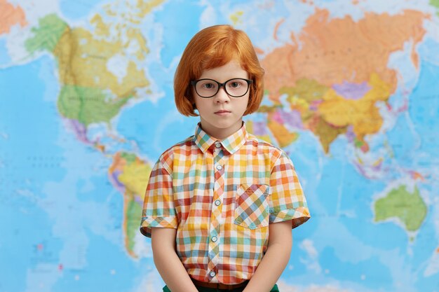 Little boy with ginger hair, wearing checkered shirt and eyeglasses, standing against map, going to school. Clever pupil standing in cabinet of geography at school, going to have lesson