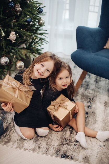 Little caucasian girls look their gifts near the christmas tree at home and smile together