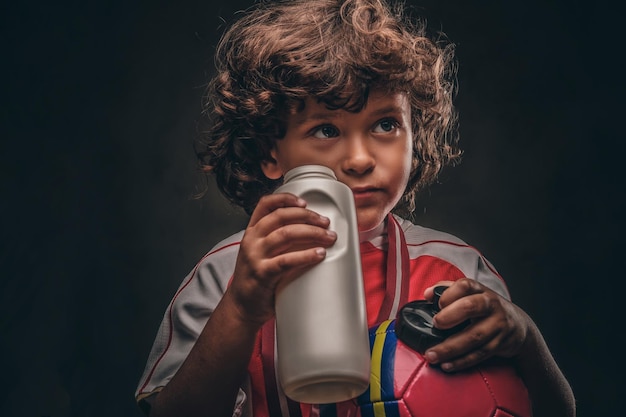 Little champion boy in sportswear holds a ball and drinking water from a bottle. Isolated on the dark textured background.