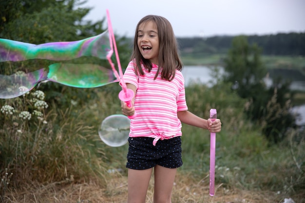 Little cheerful girl plays with big soap bubbles in nature.