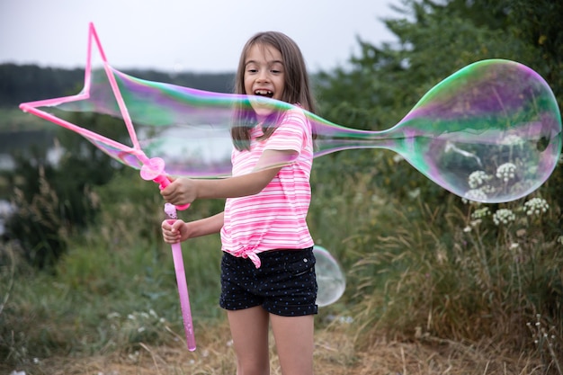 Little cheerful girl plays with big soap bubbles in nature.