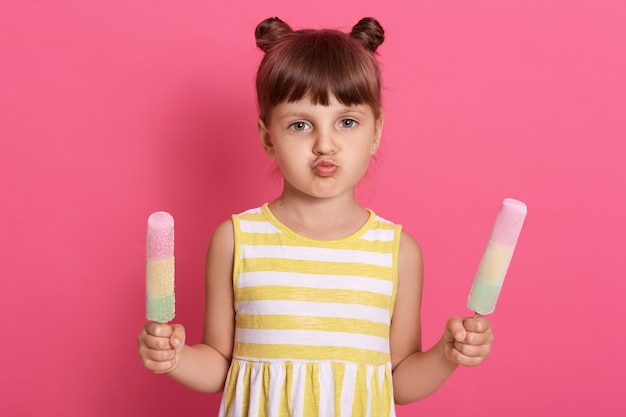 Little child girl holding ice creams in both hands,  with rounded lips, female child with two hair buns isolated over rose wall.