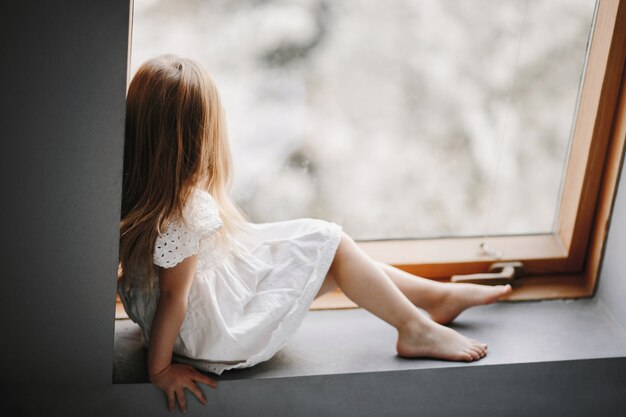 Little child in tender white dress is sitting on the windowsill