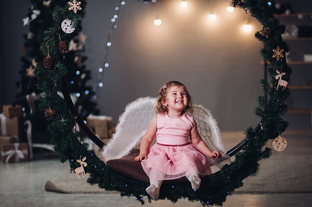 A little cute girl in a dress with angel cruces sits in a large round scenery of spruce branches