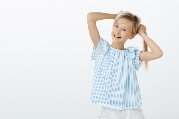 Little fashionable girl showing her first earrings to friend. Portrait of happy playful european child, holding fair hair in hands, making braids and smiling broadly, fooling around over gray wall