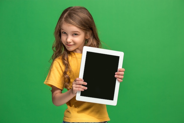 Little funny girl with tablet on green studio background. She showing something and pointing at screen.