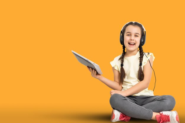 Little funny girl with tablet on yellow studio background. She showing something and pointing at screen.