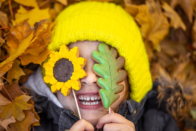 Free Photo a little funny girl in a yellow hat lies in the autumn foliage and holds gingerbread in her hands.