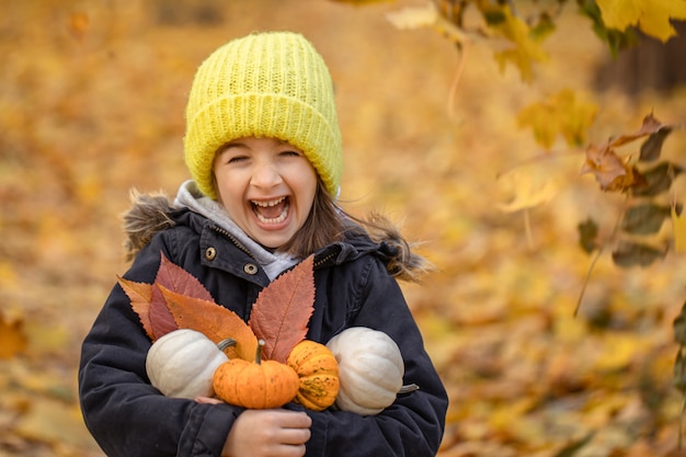 Free photo little funny girl in a yellow hat with small pumpkins in the autumn forest on a blurred background, copy space.