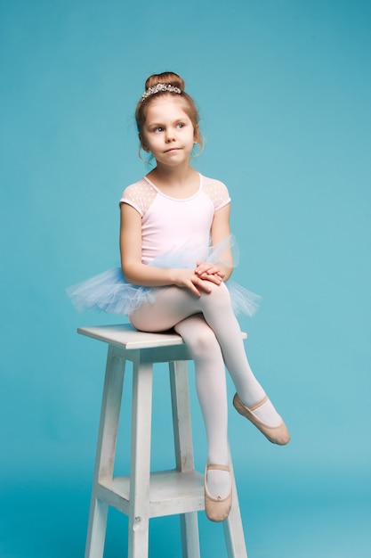 The little girl as balerina dancer sitting on white wooden chair at blue studio