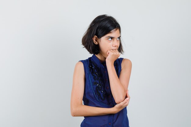 Little girl biting her fist in blue blouse and looking forgetful. front view.