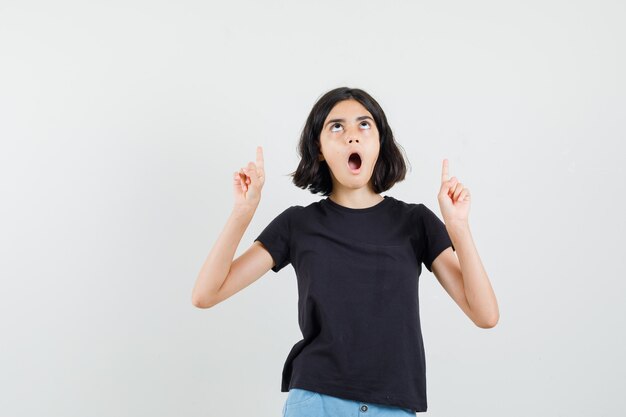 Little girl in black t-shirt, shorts pointing up and looking surprised , front view.