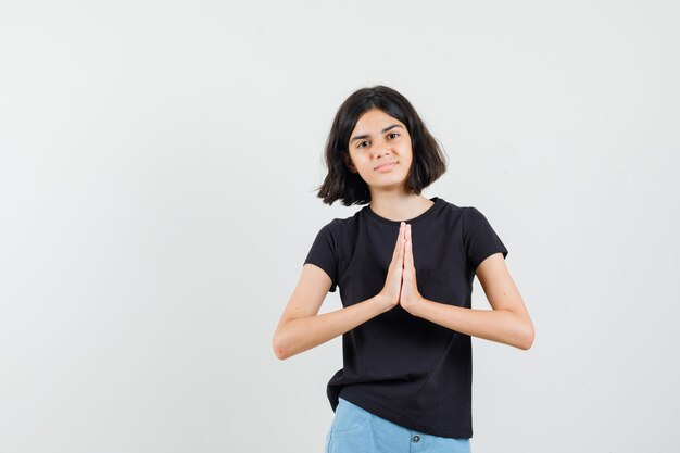 Little girl in black t-shirt, shorts showing namaste gesture and looking hopeful , front view.