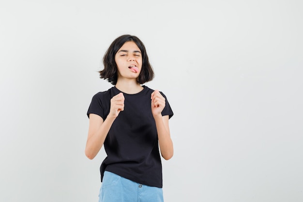 Little girl in black t-shirt, shorts sticking out tongue and looking funny , front view.