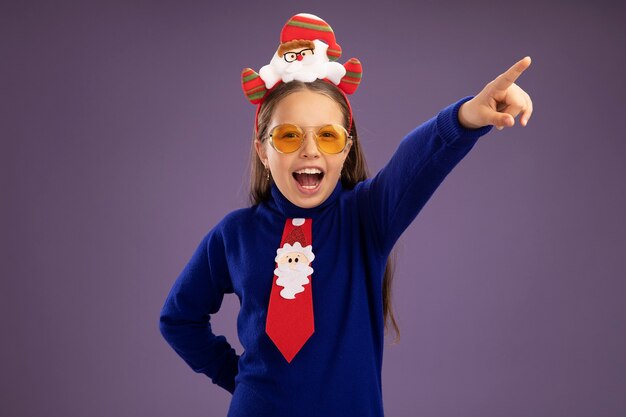 Little girl in blue turtleneck with red tie and  funny christmas rim on head happy and excited looking at camera pointing with index finger to the side standing over purple background