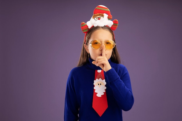 Free photo little girl in blue turtleneck with red tie and  funny christmas rim on head looking at camera making silence gesture with finger on lips  standing over purple background