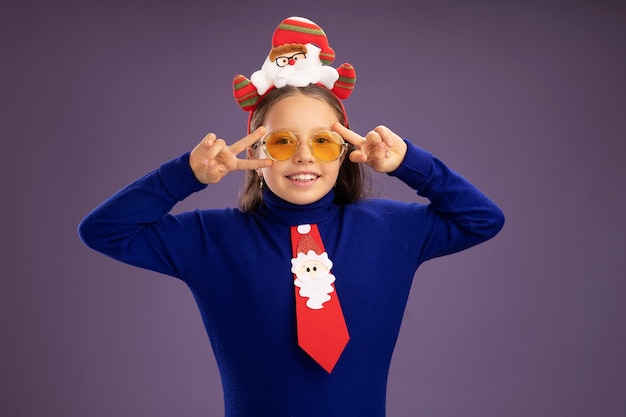 Free photo little girl in blue turtleneck with red tie and  funny christmas rim on head looking at camera smiling cheerfully showing v-sign near eyes standing over purple background